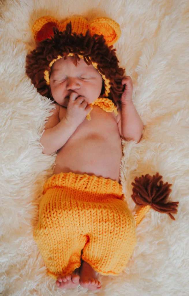 A sleeping newborn, adorned in a crocheted lion costume complete with a mane hat, rests on a soft, fluffy white surface. The vibrant orange outfit has charming brown accents on the mane and tail. The baby's hand is near their face, creating an irresistibly peaceful photo moment. © Aimee Lince Photography - Wedding photographer in Lincolnshire, Yorkshire & Nottinghamshire