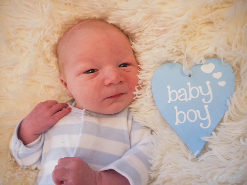 A newborn baby boy rests peacefully on a soft, fluffy cream-colored blanket. He wears a striped blue and white onesie. Beside him is a blue heart-shaped sign with "baby boy" and two small white hearts, capturing the serene moment in Cleethorpes. © Aimee Lince Photography - Wedding photographer in Lincolnshire, Yorkshire & Nottinghamshire