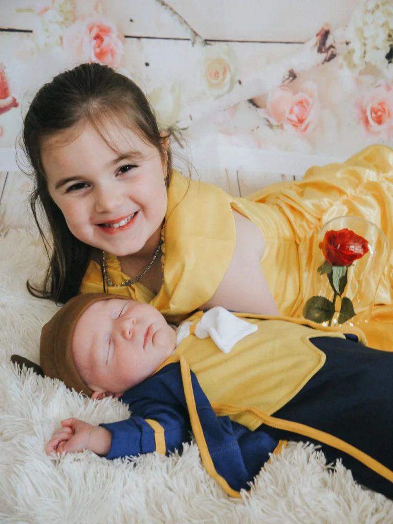 In this charming photo, a young girl in a yellow dress beams beside a sleeping baby dressed in matching yellow and blue with a brown hat. Holding a red rose, she lies on a fluffy white surface against a floral backdrop, capturing the serene essence of this Cleethorpes scene. © Aimee Lince Photography - Wedding photographer in Lincolnshire, Yorkshire & Nottinghamshire