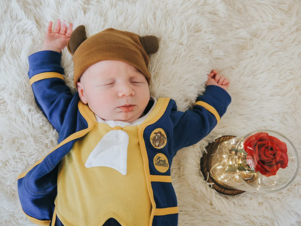 A newborn dressed in a "Beauty and the Beast" outfit lies on a fluffy white rug. The baby wears a blue jacket with gold trim, a yellow vest, a white bow tie, and a brown hat with ears. Beside the baby is a decorative glass dome containing a red rose, capturing Cleethorpes' charm. © Aimee Lince Photography - Wedding photographer in Lincolnshire, Yorkshire & Nottinghamshire