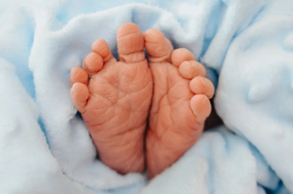 A close-up photo of a newborn's wrinkled feet gently wrapped in a soft, light blue blanket. The feet are nestled closely together, with toes slightly curled, conveying a sense of warmth and tenderness against the plush fabric. © Aimee Lince Photography - Wedding photographer in Lincolnshire, Yorkshire & Nottinghamshire