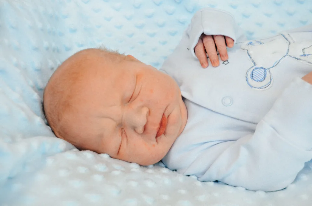 A serene photo captures a newborn baby sleeping peacefully on a light blue, textured blanket in Cleethorpes. The infant wears a light blue onesie adorned with a teddy bear design, their tiny hand resting near their face and eyes closed in tranquility. © Aimee Lince Photography - Wedding photographer in Lincolnshire, Yorkshire & Nottinghamshire