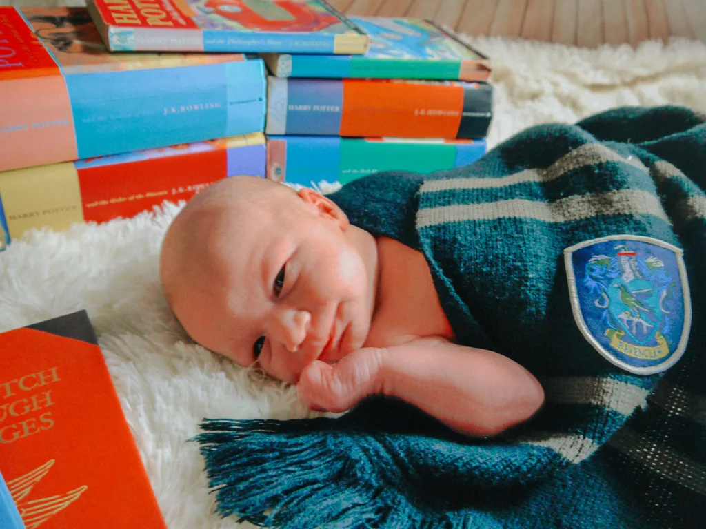 A photo captures a newborn baby wrapped in a green and blue Slytherin scarf, lying on a white fluffy surface. Nearby, a stack of colorful books adds charm to the scene. © Aimee Lince Photography - Wedding photographer in Lincolnshire, Yorkshire & Nottinghamshire
