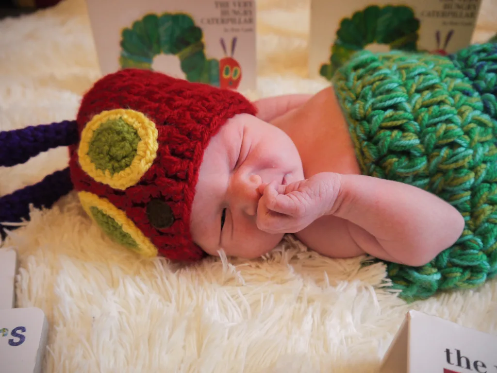 In a charming photo, a newborn baby peacefully sleeps on a soft, white fuzzy surface. They're wearing a crochet costume inspired by "The Very Hungry Caterpillar," with a red hat and matching green swaddle. The background is adorned with caterpillar-themed books, capturing the moment beautifully. © Aimee Lince Photography - Wedding photographer in Lincolnshire, Yorkshire & Nottinghamshire