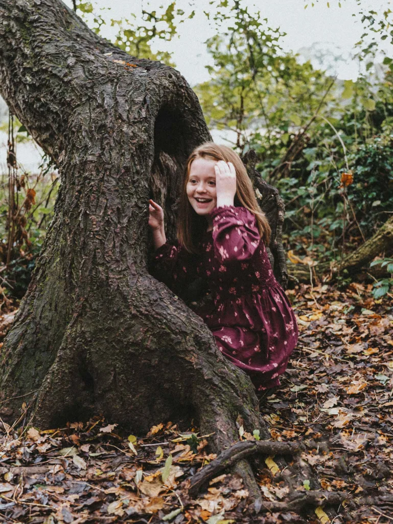 A young girl in a burgundy floral dress playfully peers out from behind the hollow of a gnarled tree. She's surrounded by fallen leaves and dense greenery, capturing a sense of adventure and nature. Perfect for a family photoshoot in Weelsby Woods, this scene is brimming with childhood wonder. © Aimee Lince Photography - Wedding photographer in Lincolnshire, Yorkshire & Nottinghamshire