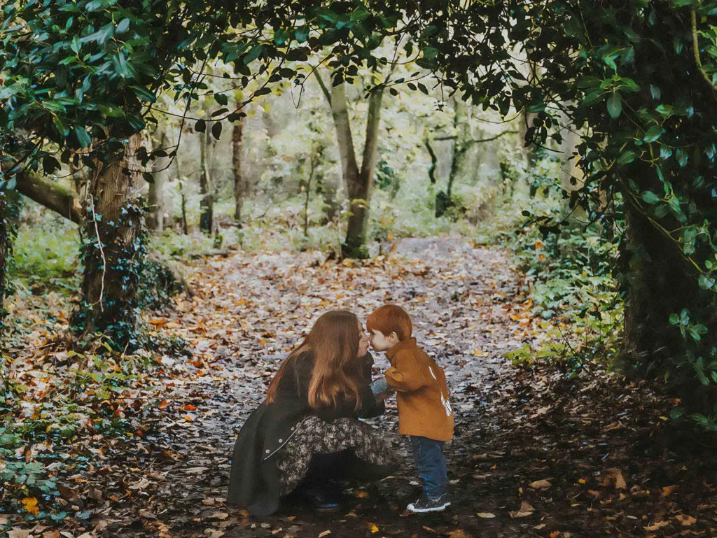 A woman with long hair kneels on a leaf-covered forest path in Weelsby Woods, facing a young child in a brown coat. The child leans in, lips puckered for a kiss. The surrounding trees form a natural arch, capturing the essence of Stacey & Her Kids during their serene family photoshoot. © Aimee Lince Photography - Wedding photographer in Lincolnshire, Yorkshire & Nottinghamshire