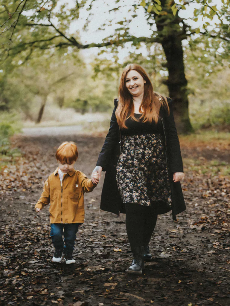 Stacey and her kids stroll hand in hand through Weelsby Woods, where a woman in a black coat and floral dress walks beside a young child clad in a mustard jacket. The leafy dirt path is adorned with fallen leaves, and the lush trees create the perfect backdrop for this spontaneous family photoshoot. © Aimee Lince Photography - Wedding photographer in Lincolnshire, Yorkshire & Nottinghamshire