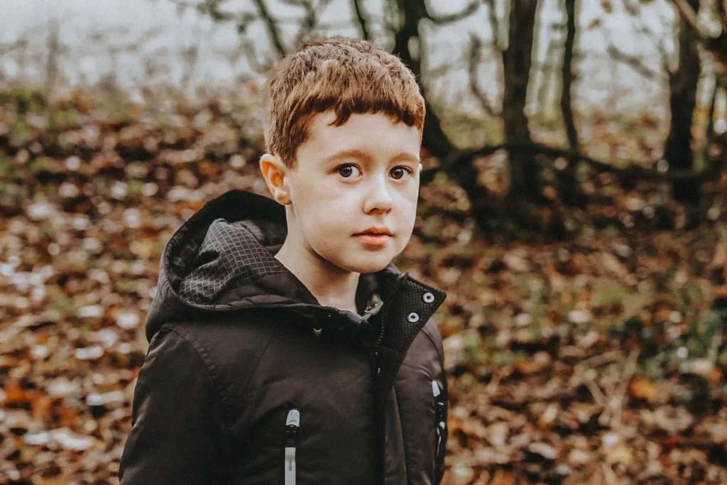 Amid the tranquil setting of Weelsby Woods, a boy with short brown hair and a serious expression stands outdoors, wearing a dark jacket with a hood. The forest floor is blanketed in fallen leaves, capturing the essence of autumn during Stacey & Her Kids' family photoshoot. © Aimee Lince Photography - Wedding photographer in Lincolnshire, Yorkshire & Nottinghamshire