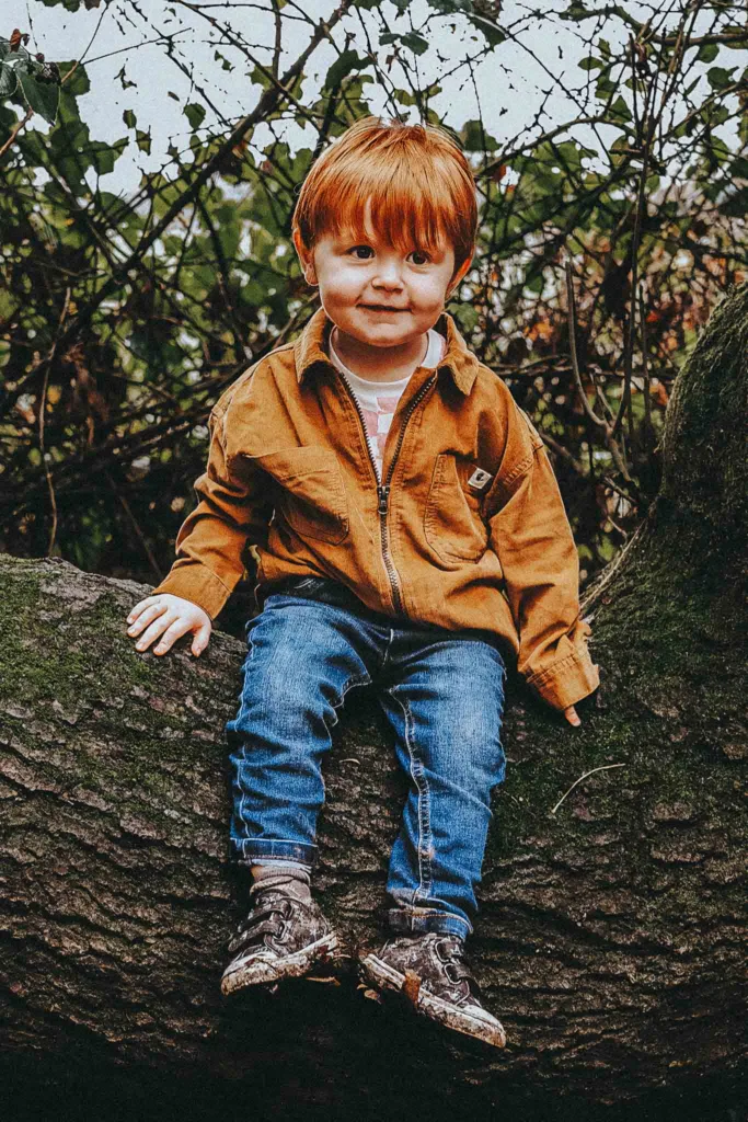 A red-haired toddler sits on a large tree trunk during a family photoshoot at Weelsby Woods, wearing a brown jacket, blue jeans, and patterned sneakers. He has a playful expression and slightly tilts his head amid the dense, leafy foliage. © Aimee Lince Photography - Wedding photographer in Lincolnshire, Yorkshire & Nottinghamshire