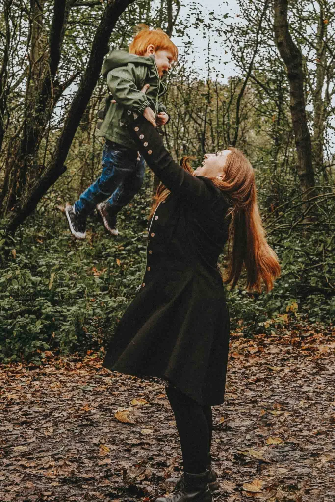 In Weelsby Woods, Stacey joyfully lifts her child, adorned with red hair and a green jacket. Surrounded by sparse trees and fallen leaves, the scene captures a cherished family photoshoot moment. Both appear happy, savoring their playful adventure in the heart of autumn. © Aimee Lince Photography - Wedding photographer in Lincolnshire, Yorkshire & Nottinghamshire