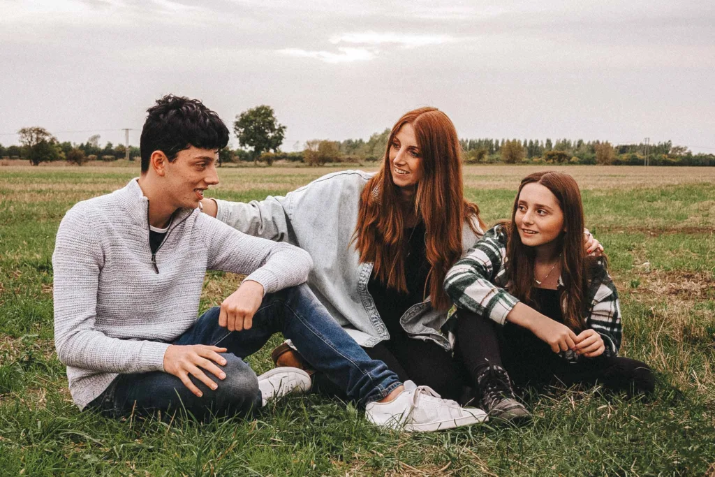 In Weelsby Woods, a family photoshoot comes to life as three people sit on the grass in conversation. A young man in a gray sweater and jeans sits on the left, Kelly with long red hair wears a denim jacket, and a girl in plaid completes the scene. The overcast sky adds an air of intimacy. © Aimee Lince Photography - Wedding photographer in Lincolnshire, Yorkshire & Nottinghamshire