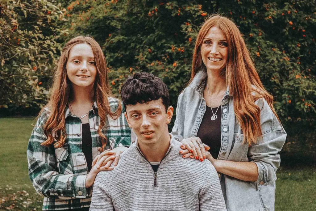 A woman with long red hair stands smiling alongside a young girl with similar hair and a young boy during a family photoshoot. The girl wears a green plaid shirt, and the boy wears a light gray sweater. They are outdoors in Weelsby Woods, surrounded by lush green foliage as she places her hands on the boy's shoulders. © Aimee Lince Photography - Wedding photographer in Lincolnshire, Yorkshire & Nottinghamshire