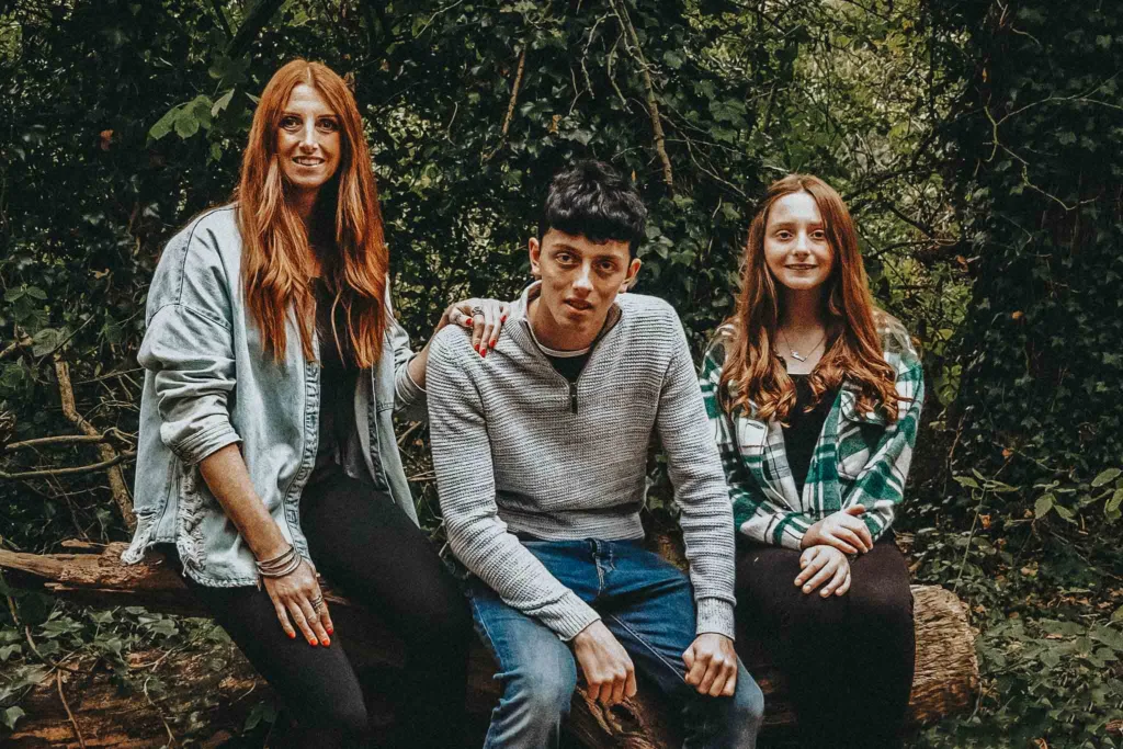 In Weelsby Woods, a serene setting perfect for a family photoshoot, Kelly and her teens sit on a fallen tree. On the left, with long red hair and a denim jacket; in the middle, dark-haired with a gray sweater; and on the right, another redhead sporting a green plaid shirt—all enveloped by lush greenery. © Aimee Lince Photography - Wedding photographer in Lincolnshire, Yorkshire & Nottinghamshire