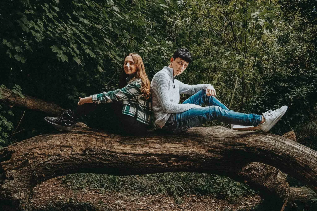 A young woman and man sit back-to-back on a large tree branch in Weelsby Woods. The woman sports a green flannel shirt and black boots; the man wears a gray sweater, jeans, and white sneakers. Surrounded by lush foliage, Kelly & Her Teens capture a relaxed and adventurous family photoshoot moment. © Aimee Lince Photography - Wedding photographer in Lincolnshire, Yorkshire & Nottinghamshire