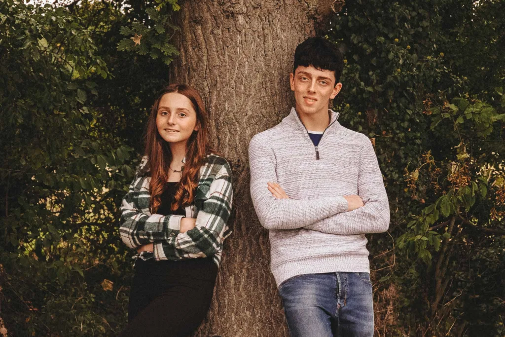 Two teens stand outdoors against a tree trunk in Weelsby Woods, surrounded by lush green foliage. The one on the left, with long hair, sports a green plaid shirt and black pants. The one on the right wears a light gray sweater and blue jeans. Both have their arms crossed and smile slightly during the family photoshoot. © Aimee Lince Photography - Wedding photographer in Lincolnshire, Yorkshire & Nottinghamshire