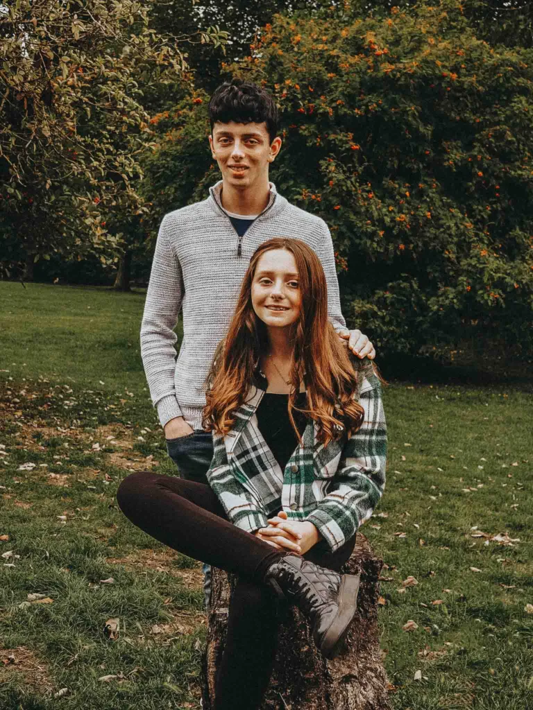 In Weelsby Woods, a young woman with long brown hair, wearing a green plaid shirt and black leggings, sits on a tree stump. A young man in a gray sweater and jeans stands behind her, one hand resting on her shoulder. Green grass and bushes with orange berries create the perfect backdrop for this family photoshoot. © Aimee Lince Photography - Wedding photographer in Lincolnshire, Yorkshire & Nottinghamshire