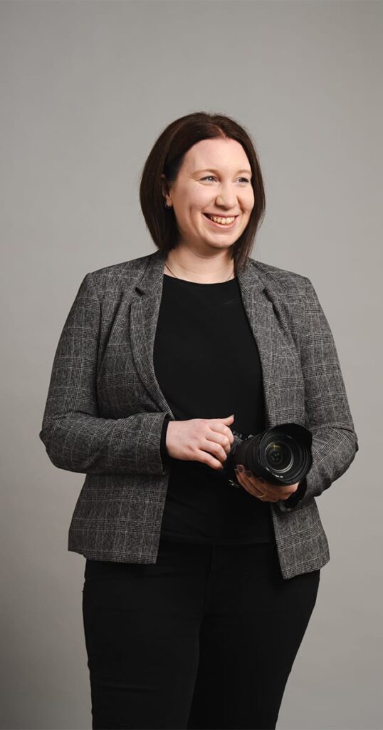 A smiling person with shoulder-length brown hair holds a camera lens, ready to capture moments for my blog. They are wearing a gray plaid blazer over a black shirt and black pants, set against a neutral gray background. © Aimee Lince Photography