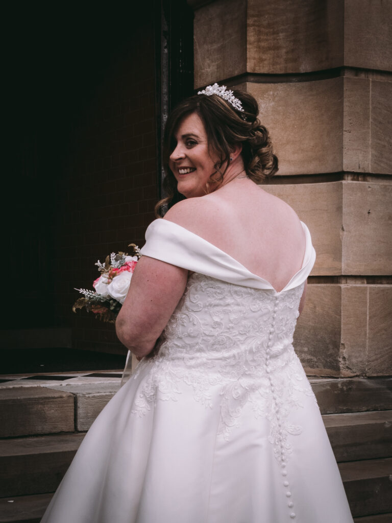 A bride wearing an off-shoulder white wedding gown stands smiling, looking back over her shoulder. She holds a bouquet of flowers in her left hand and wears a tiara. The background features Cleethorpes' light brown stone architectural details and a dark entrance. © Aimee Lince Photography