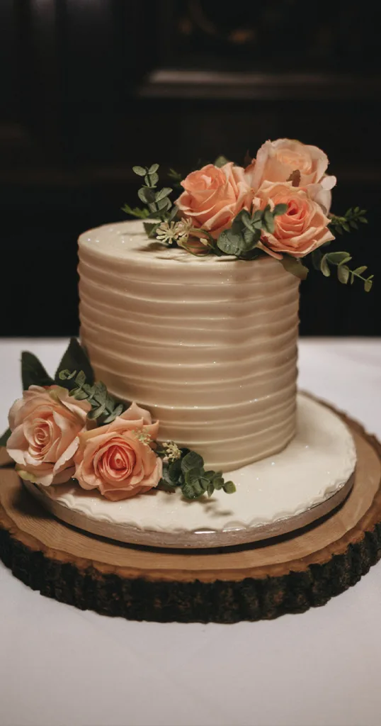 A two-tiered white cake with ribbed icing is adorned with peach roses and greenery, offering a budget-friendly elegance. It rests on a wooden slab with a white base layer atop a white tablecloth. The flowers cascade gracefully down the side, adding a rustic and romantic touch. © Aimee Lince Photography - Wedding photographer in Lincolnshire, Yorkshire & Nottinghamshire