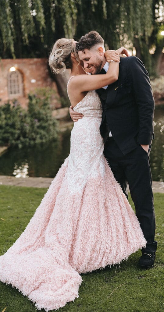 A bride in a pink, textured gown with lace detail embraces a groom in a black suit by the pond at Stallingborough Grange Hotel. Surrounded by trees and a brick wall, the scene captures the joy and romance of their wedding day. © Aimee Lince Photography