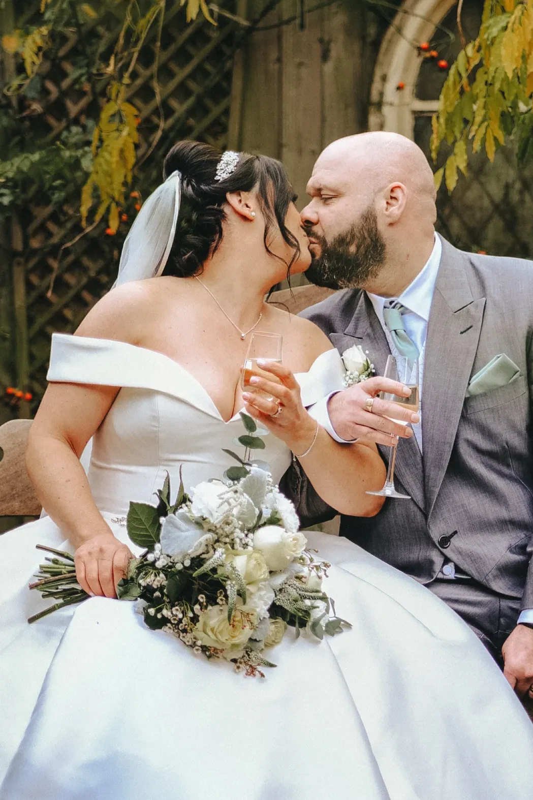 A newlywed couple shares a tender kiss on a wooden bench, the bride holding a bouquet, with a backdrop of autumn leaves and a rustic fence, symbolizing their wedding union amidst the serene garden setting