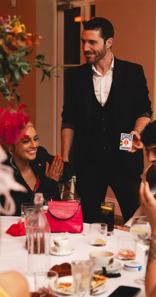 A man in a black suit and white shirt dazzles guests with a card trick at a table, offering unique wedding entertainment. A smiling woman sporting a large pink flower headpiece applauds. Drinks, a red purse, and plates of food adorn the table as other guests and flowers fill the joyful scene. © Aimee Lince Photography - Wedding photographer in Lincolnshire, Yorkshire & Nottinghamshire