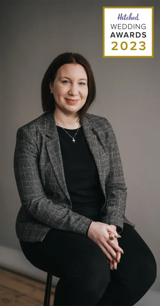 A person with shoulder-length dark hair sits against a gray background, wearing a gray plaid blazer over a black top. They smile slightly, hands clasped in their lap. The prestigious "Hitched Wedding Awards 2023" appears at the top right in a white and gold-bordered box, highlighting their achievement. © Aimee Lince Photography - Wedding photographer in Lincolnshire, Yorkshire & Nottinghamshire