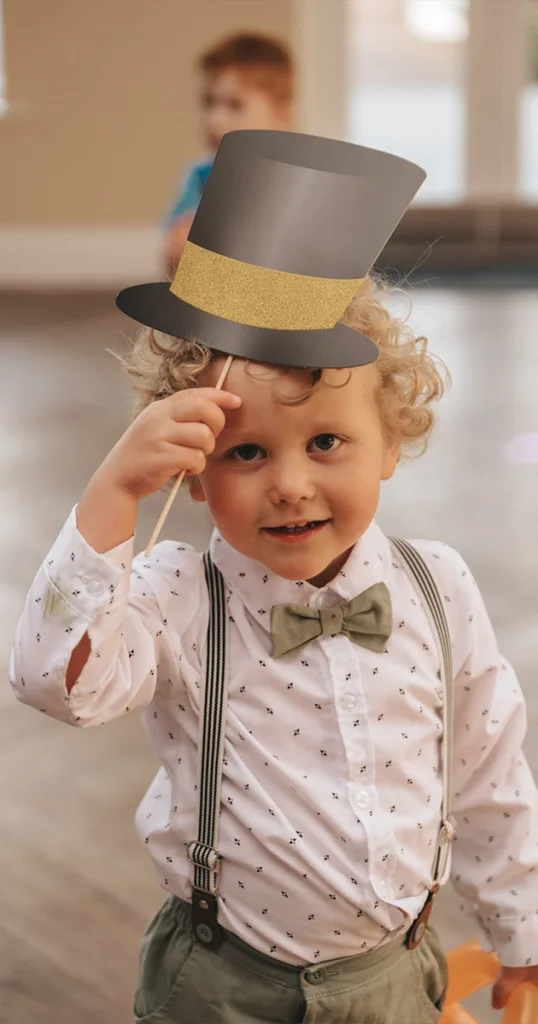 A young child with curly blonde hair holds up a paper top hat on a stick, offering charming wedding entertainment ideas. He wears a white shirt with small black patterns, a gray bow tie, and gray suspenders. The background shows a blurred indoor setting with another child playing in the distance. © Aimee Lince Photography - Wedding photographer in Lincolnshire, Yorkshire & Nottinghamshire