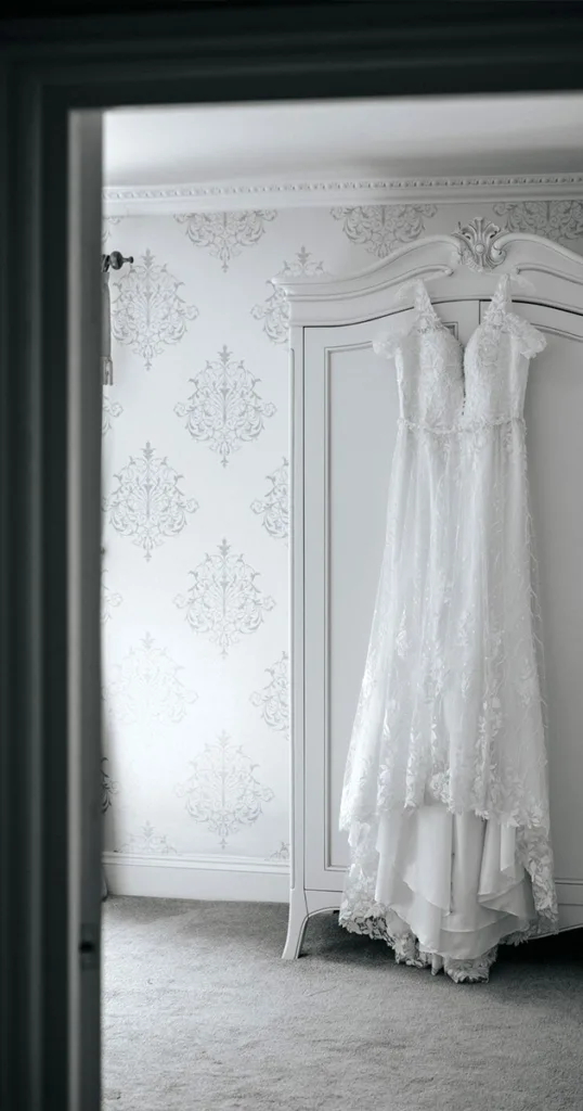 A white lace wedding dress, embodying the essence of wedding blues, hangs on an ornate white armoire in a softly lit room. Light gray carpet and floral-patterned wallpaper craft an elegant and serene atmosphere, while the door frame in the foreground beautifully frames this romantic scene. © Aimee Lince Photography - Wedding photographer in Lincolnshire, Yorkshire & Nottinghamshire