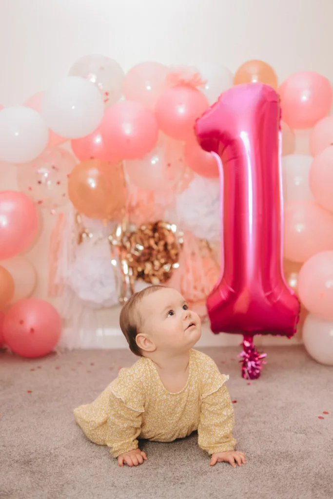 A baby in a yellow patterned outfit crawls on a gray carpet. Behind her, a large pink foil balloon shaped like the number one is surrounded by pink, white, and gold balloons, capturing the essence of cake smash photography. Confetti is scattered around, enhancing the festive atmosphere as she looks upward with curiosity. © Aimee Lince Photography - Wedding photographer in Lincolnshire, Yorkshire & Nottinghamshire