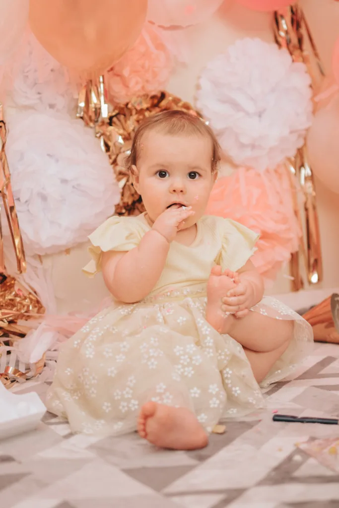 A baby in a light yellow dress with ruffled sleeves sits on a patterned blanket, resembling an adorable scene from a photography session. The little one nibbles a finger while holding a foot, surrounded by pink, white, and gold decorative paper flowers and balloons—perfect for capturing precious cake smash moments. © Aimee Lince Photography - Wedding photographer in Lincolnshire, Yorkshire & Nottinghamshire