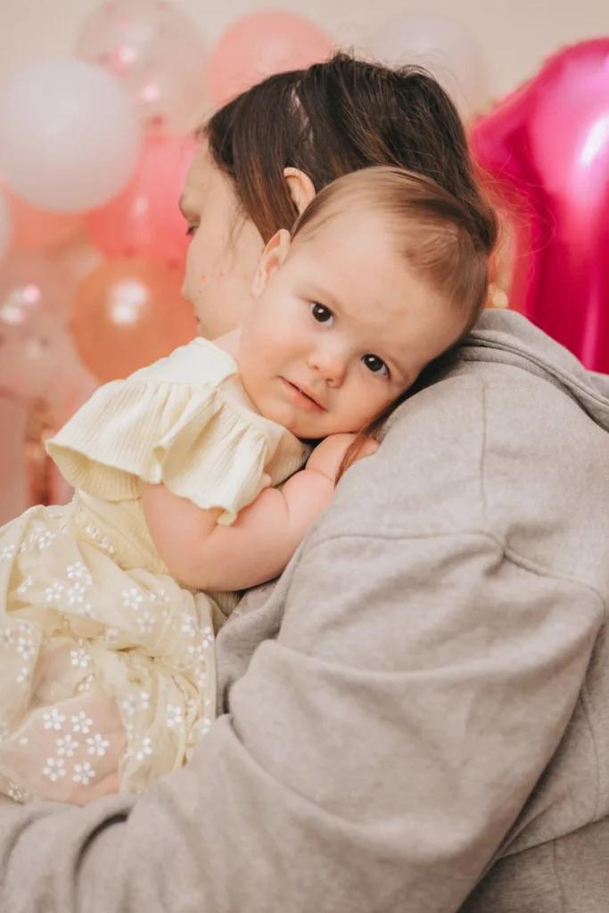 A baby in a cream dress adorned with floral patterns rests its head on a shoulder clad in a gray hoodie. The background, dotted with pink and white balloons, sets the stage for what feels like a serene moment captured by photography amid festive preparations for a cake smash celebration. © Aimee Lince Photography - Wedding photographer in Lincolnshire, Yorkshire & Nottinghamshire