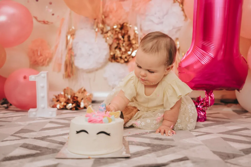 A baby in a light yellow dress eagerly leans forward on a patterned floor, preparing to smash the cake adorned with icing and a unicorn figure. Pink and peach balloons, a large pink number 1 balloon, and a white lit number 1 complete the festive photography backdrop for this charming first birthday celebration. © Aimee Lince Photography - Wedding photographer in Lincolnshire, Yorkshire & Nottinghamshire
