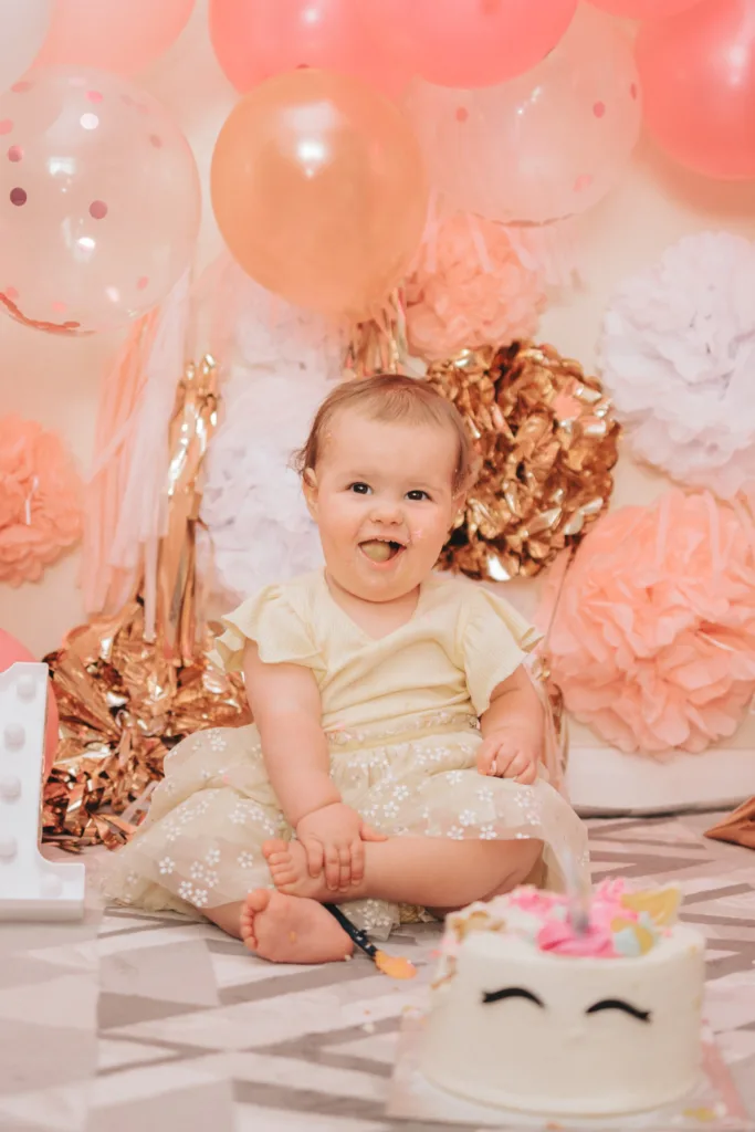 A joyful baby in a cream dress sits barefoot on the floor, playfully engaging in a cake smash. The background features pink, peach, and gold balloons with matching paper decorations. A decorative white number one and a cake with a smiling face design add to the cheerful photography setting. © Aimee Lince Photography - Wedding photographer in Lincolnshire, Yorkshire & Nottinghamshire