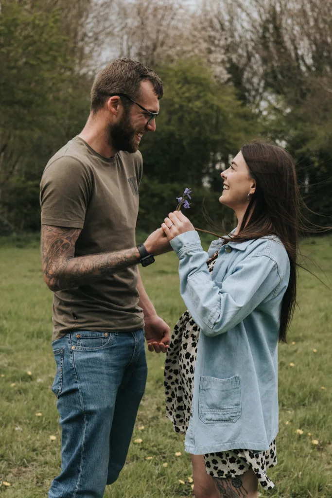 A man and woman stand in a grassy field at Hall Farm. The man, wearing a brown shirt and jeans, smiles while holding a single flower. The woman, in a polka dot dress and denim jacket, smiles at him with windblown hair as trees and a cloudy sky form the background. © Aimee Lince Photography - Wedding photographer in Lincolnshire, Yorkshire & Nottinghamshire