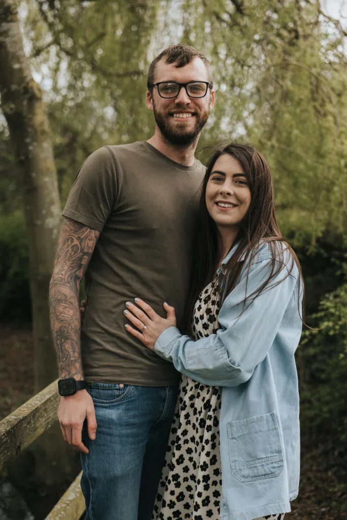 A man with glasses and tattoos on his right arm stands on a wooden bridge at Hall Farm, smiling with his arm around a woman. The woman, also smiling, wears a polka-dot dress with a blue jacket. They are outdoors, surrounded by trees and greenery in the background. © Aimee Lince Photography - Wedding photographer in Lincolnshire, Yorkshire & Nottinghamshire