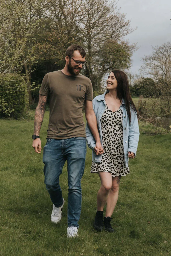 A couple strolls hand in hand across a hall farm's grassy field. The man, in sunglasses, a brown t-shirt, and jeans, gazes at the woman in her black and white polka dot dress with a denim jacket. Both beam joyfully, surrounded by trees under a cloudy sky. © Aimee Lince Photography - Wedding photographer in Lincolnshire, Yorkshire & Nottinghamshire