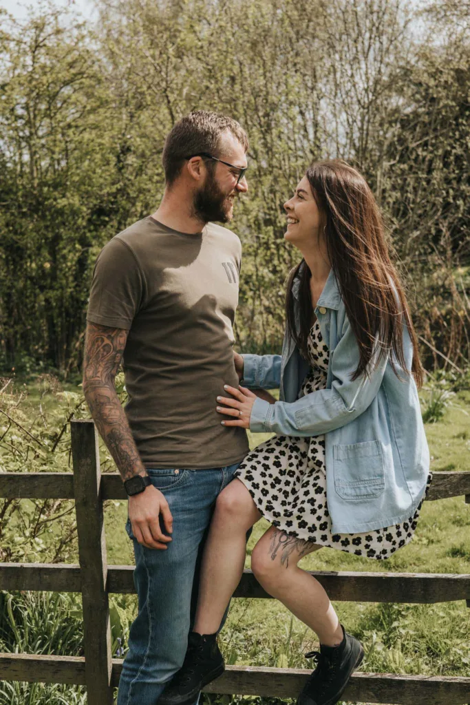 A bearded man in a green t-shirt and jeans stands beside a smiling woman sitting on a fence at Hall Farm. The woman is wearing a black and white patterned dress and denim jacket. They are outdoors, surrounded by greenery, and appear to be enjoying a sunny day. © Aimee Lince Photography - Wedding photographer in Lincolnshire, Yorkshire & Nottinghamshire