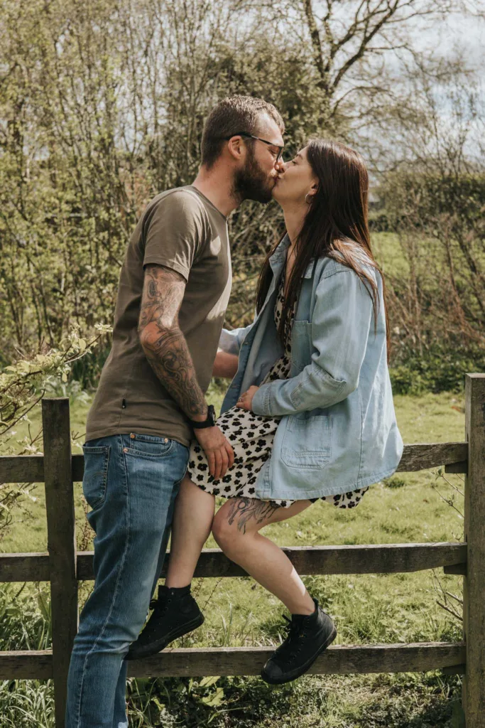 A couple shares a kiss by a wooden fence on Hall Farm. The man, adorned with tattoos and glasses, wears a brown t-shirt and jeans as he leans forward. The woman, in a patterned dress, denim jacket, and black boots, sits on the fence with trees and an open field stretching beyond them. © Aimee Lince Photography - Wedding photographer in Lincolnshire, Yorkshire & Nottinghamshire