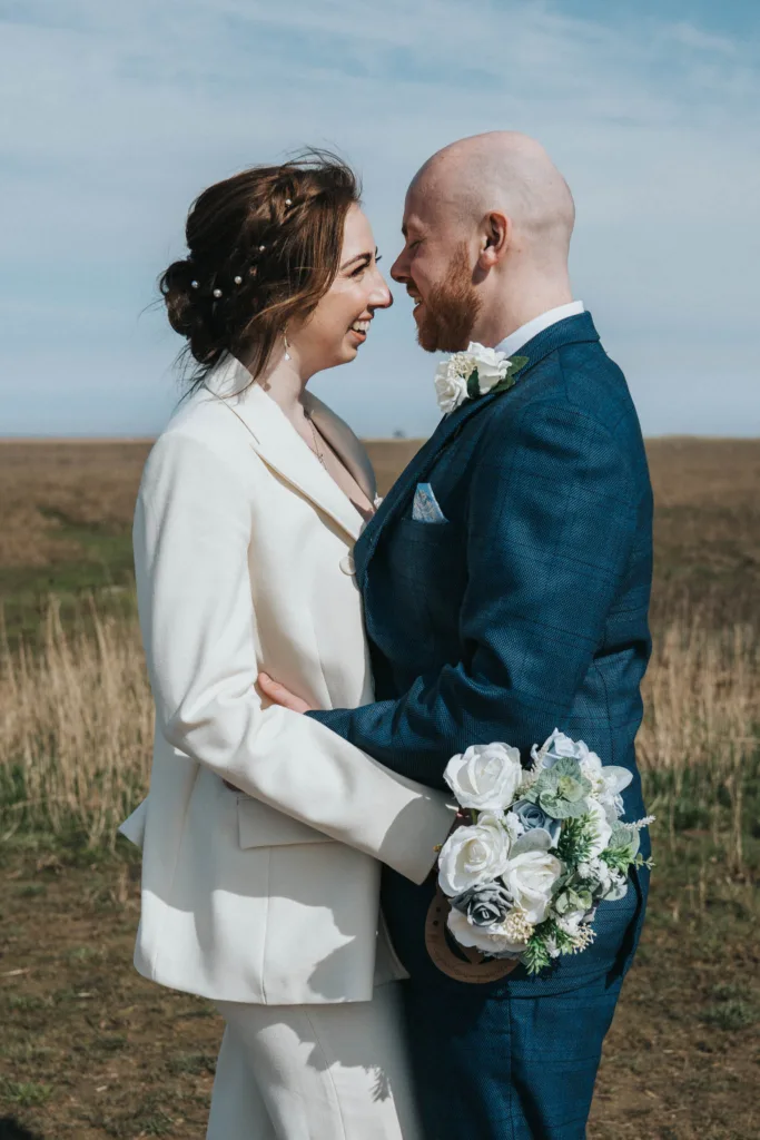 A couple stands embracing outdoors, smiling at each other under the clear blue sky at Cleethorpes seafront. The woman wears a white suit with delicate floral details in her hair, while the man, in a blue suit, holds a bouquet with white roses—a perfect scene for wedding photography. © Aimee Lince Photography - Wedding photographer in Lincolnshire, Yorkshire & Nottinghamshire