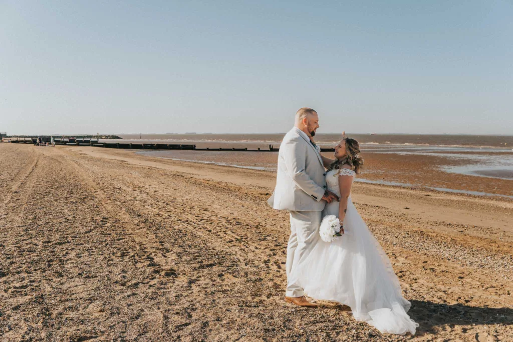 A bride in a white gown and groom in a light gray suit stand on Cleethorpes seafront. They gaze at each other lovingly, with the ocean and clear blue sky in the background. The bride holds a bouquet of white flowers, as their footprints mark the sandy beach—a perfect moment for wedding photography. © Aimee Lince Photography - Wedding photographer in Lincolnshire, Yorkshire & Nottinghamshire
