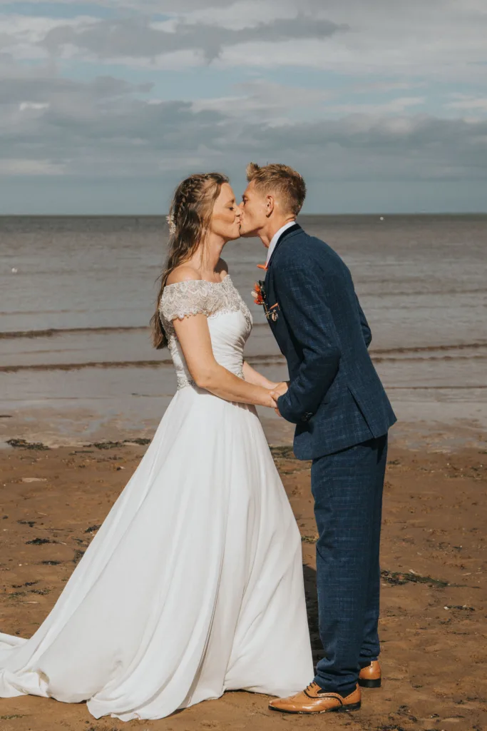 A bride and groom kiss on the sandy Cleethorpes seafront. The bride wears a white dress with lace off-shoulder sleeves, while the groom is in a blue suit with tan shoes. They hold hands against a backdrop of calm sea under a partly cloudy sky—perfect for wedding photography. © Aimee Lince Photography - Wedding photographer in Lincolnshire, Yorkshire & Nottinghamshire