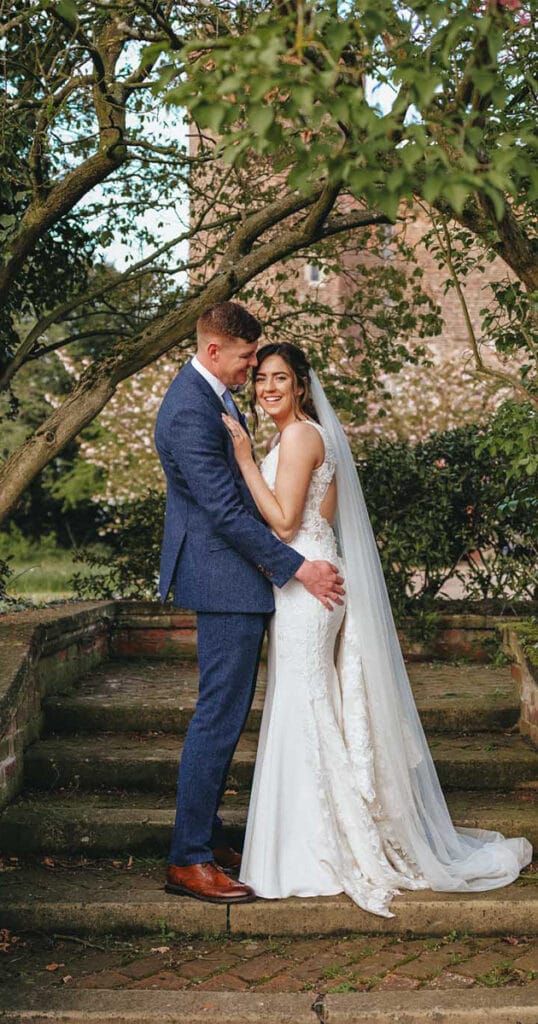 A couple in wedding attire stands on stone steps surrounded by greenery at Hodsock Priory. The groom dons a blue suit and brown shoes, while the bride wears a white lace gown and veil. They hug and smile, captured beautifully by photography, with tall trees and a brick building in the background. © Aimee Lince Photography