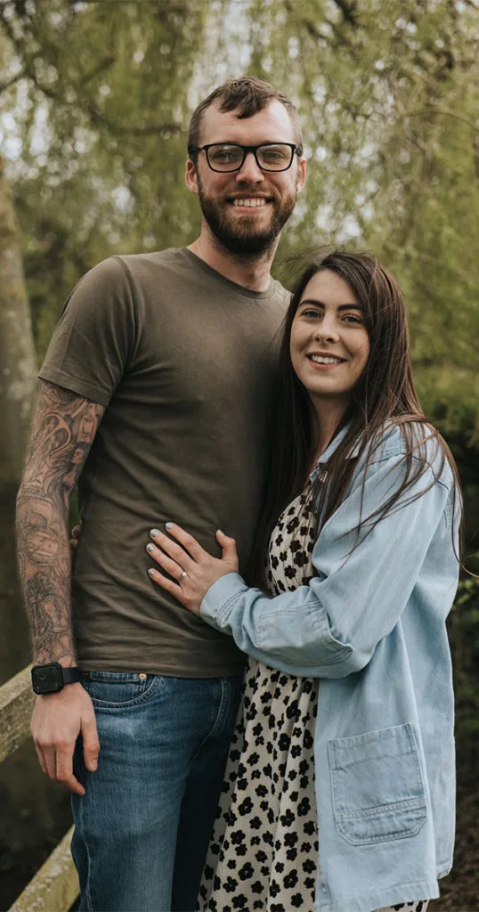 A couple stands on a wooden bridge at Hall Farm, smiling. The man wears glasses, a brown t-shirt, and jeans, showing tattooed arms. The woman wears a floral dress and denim jacket, long dark hair flowing. Her hand on his chest highlights an engagement ring. Green trees surround them in the background. © Aimee Lince Photography - Wedding photographer in Lincolnshire, Yorkshire & Nottinghamshire