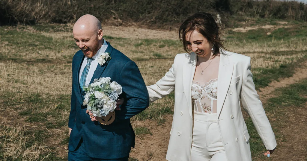 A couple smiles widely as they walk outdoors on a grassy path, embodying the essence of wedding photography. The man, in a blue suit, holds a white rose bouquet. The woman, wearing a white lace top with a matching blazer and pants, gently holds his arm. The sky is clear, embracing the charm of Cleethorpes seafront in the background. © Aimee Lince Photography - Wedding photographer in Lincolnshire, Yorkshire & Nottinghamshire