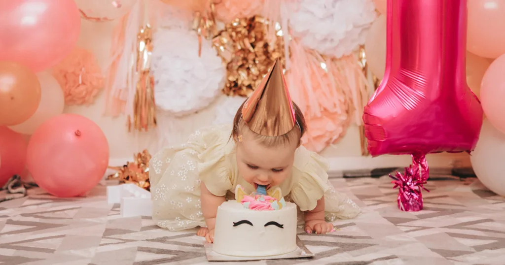 A baby wearing a gold party hat and beige dress leans forward to smash a cake on the floor, captured in charming photography. The pastel-colored frosting features closed-eye decorations. Pink and peach balloons surround the scene, with a large pink number one balloon hinting at a first birthday celebration. © Aimee Lince Photography - Wedding photographer in Lincolnshire, Yorkshire & Nottinghamshire