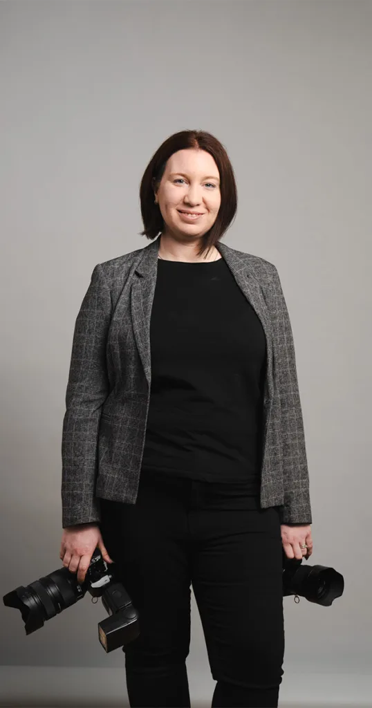 A person with shoulder-length brown hair stands against a sleek gray background, exuding a hybrid style in a gray plaid blazer over a black shirt and pants. With a camera in each hand, they flash a subtle smile. © Aimee Lince Photography - Wedding photographer in Lincolnshire, Yorkshire & Nottinghamshire