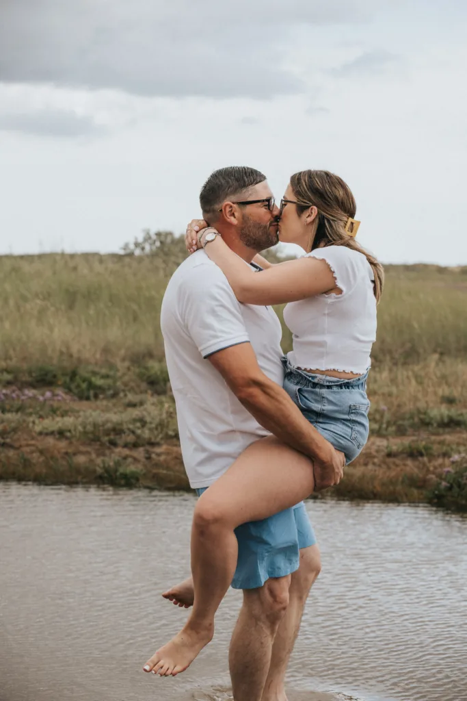 A couple kisses by a shallow body of water at Cleethorpes beach. The man, in a white t-shirt and blue shorts, holds the woman, dressed in a white top and denim shorts. She wraps her legs around his waist, as grassy terrain unfolds under a cloudy sky. © Aimee Lince Photography - Wedding photographer in Lincolnshire, Yorkshire & Nottinghamshire