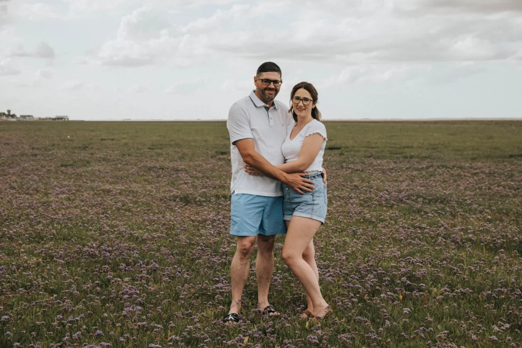 A couple stands in a field of purple flowers under a cloudy sky, reminiscent of Cleethorpes Beach. Both wear glasses and summer attire: the man in a white shirt and blue shorts, the woman in a white top and denim shorts. They smile and embrace, surrounded by a vast, open landscape. © Aimee Lince Photography - Wedding photographer in Lincolnshire, Yorkshire & Nottinghamshire