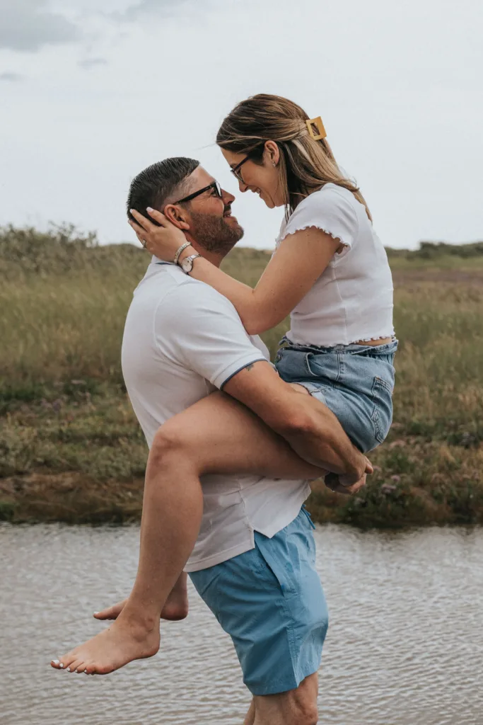 A man in a white t-shirt and blue shorts embraces a woman wearing a white crop top and denim shorts. She sits on the man's knee with her legs wrapped around his waist. They smile at each other near Cleethorpes beach, with the grassy field leading to water under a cloudy sky. © Aimee Lince Photography - Wedding photographer in Lincolnshire, Yorkshire & Nottinghamshire